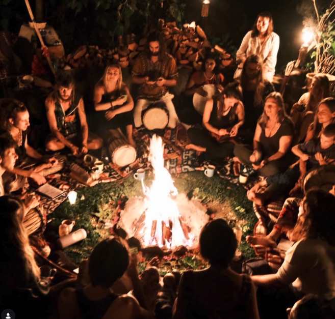 People gathered in a circle around an evening fire, sharing songs and drums